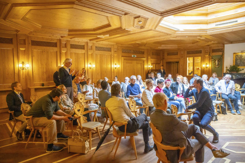 Eine Gruppe von Menschen sitzt in einem holzvertäfelten Raum und nimmt an einer angeregten Diskussion oder Veranstaltung teil – gemütliche, alpine Atmosphäre.